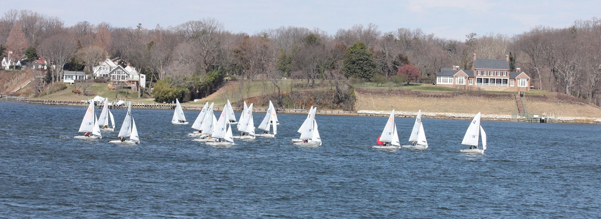 A group of small sailboats with white sails race across a wide river, with large houses and leafless trees visible on the opposite bank under a partly cloudy sky.