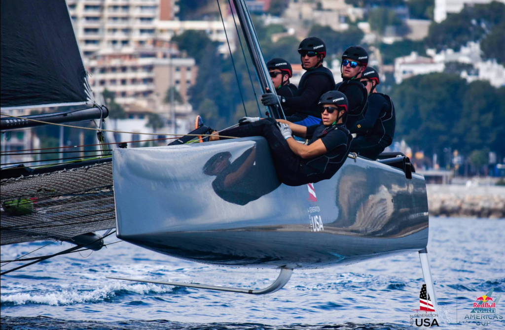 A sailing team in black gear and helmets leans out over the edge of a sleek racing boat above the water, with city buildings and trees in the blurry background. Logos and an American flag are visible on the boat’s hull.
