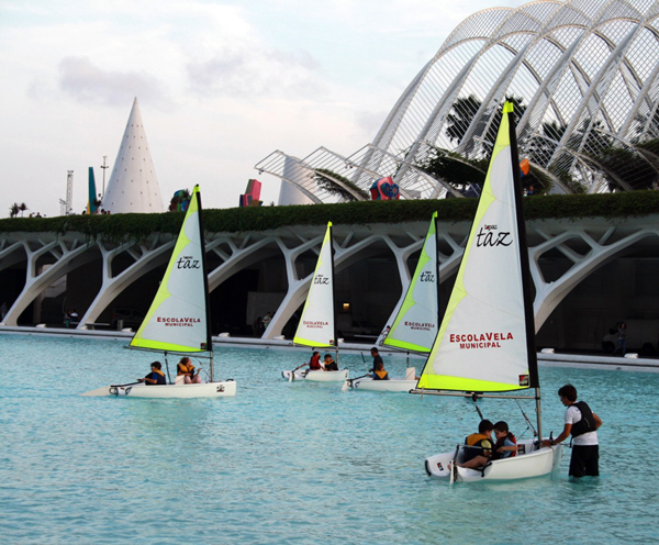 Several small sailboats with children and instructors are sailing in a turquoise pool, with modern white architectural structures and greenery in the background.