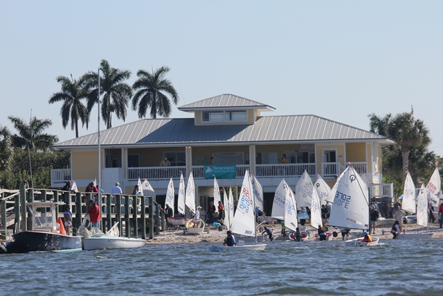 A group of small sailboats and people gather on a sandy shore in front of a yellow building with palm trees and a dock, seen from the water on a sunny day.