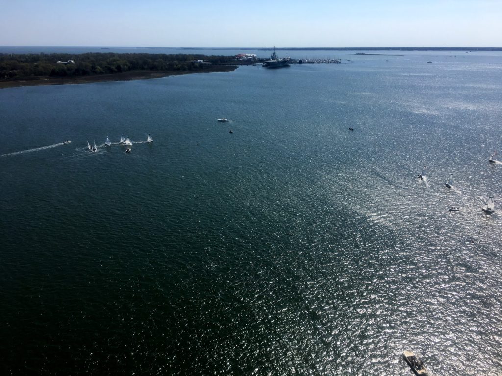 A wide view of a calm, sunlit body of water with several small boats scattered across the surface. In the distance, there is a forested shoreline and a few buildings near the water’s edge under a clear blue sky.