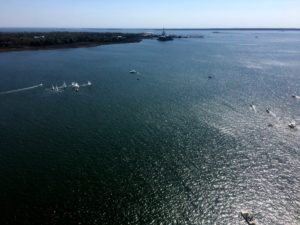 A wide view of a calm, sunlit body of water with several small boats scattered across the surface. In the distance, there is a forested shoreline and a few buildings near the water’s edge under a clear blue sky.