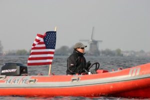 A person in a black jacket and cap drives an orange inflatable boat with an American flag on the water. A windmill is visible in the background under a clear sky.