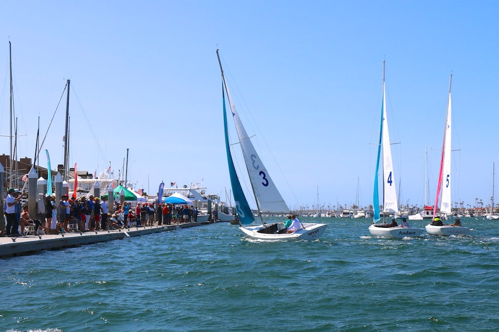 Small sailboats with numbered sails race near a marina as spectators watch from a dock lined with tents on a sunny day, with choppy blue water in the foreground.