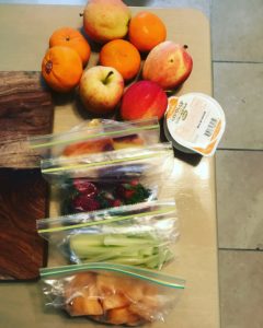A tabletop with apples, oranges, a cup of mandarin oranges, and three ziplock bags containing cut fruit, celery sticks, and sliced strawberries.