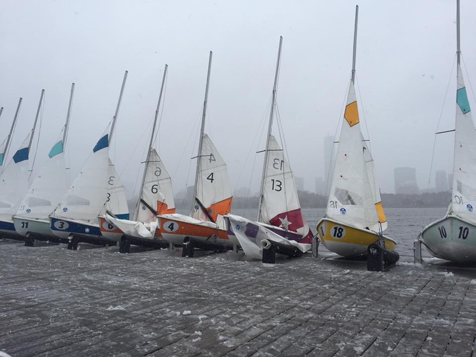 Several small sailboats with colorful numbers and markings are lined up on a snowy dock, with a foggy city skyline visible in the background.