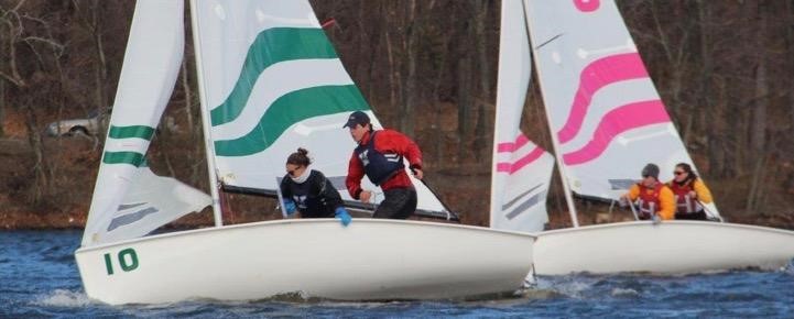 Two small sailboats with crews of two race on a lake; the first boat has green and white sails and the second has pink and white sails. People wear life vests and trees line the shore in the background.