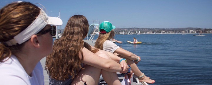 Three people sit on a dock by the water, looking out at kayakers and paddleboarders on a sunny day. All three wear casual clothing and hats, and the background shows distant buildings and hills.