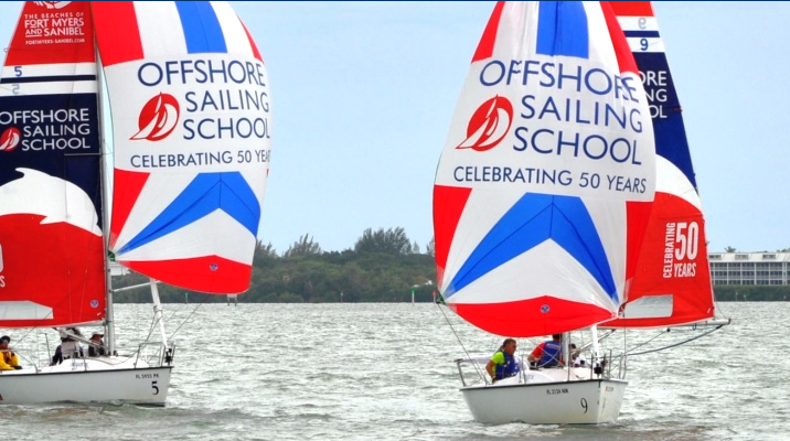 Two sailboats with colorful sails that read “Offshore Sailing School Celebrating 50 Years” glide on the water during a sailing event, with trees and buildings visible in the background.