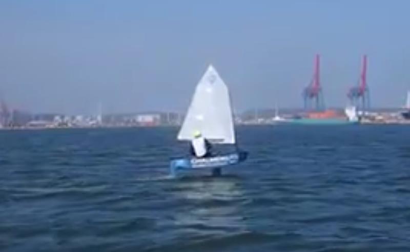 A person sails a small boat with a white sail on a body of water, with cranes and industrial structures visible in the background under a clear blue sky.