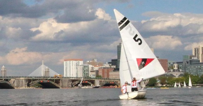 Two people sail a small white sailboat with the number 5 and an 'H' logo on its sail, on a river with a city skyline, bridge, and partly cloudy sky in the background.