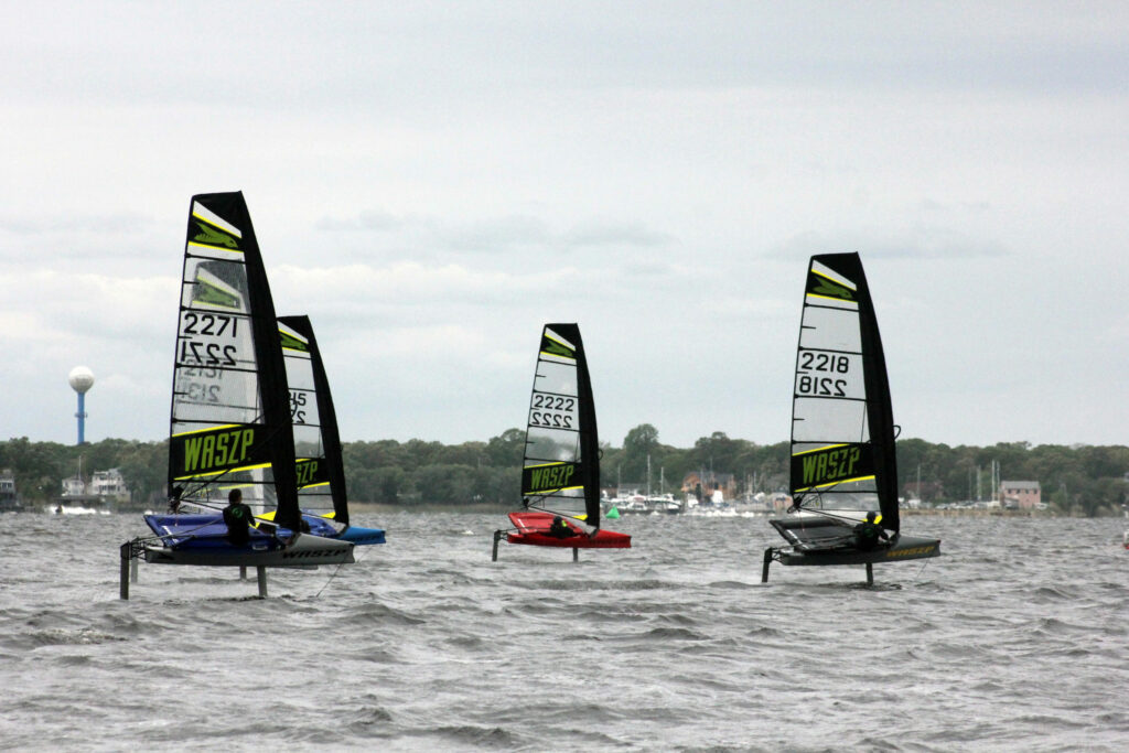 Four small sailboats with tall sails and hydrofoils race on choppy water under a cloudy sky, with trees, houses, and a water tower visible in the background.