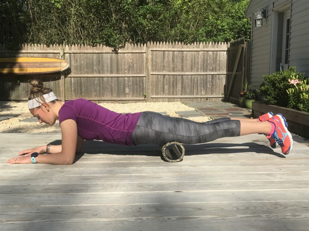 A woman in athletic wear uses a foam roller under her thighs while in a plank position on a wooden deck outdoors, with a fence and greenery in the background.