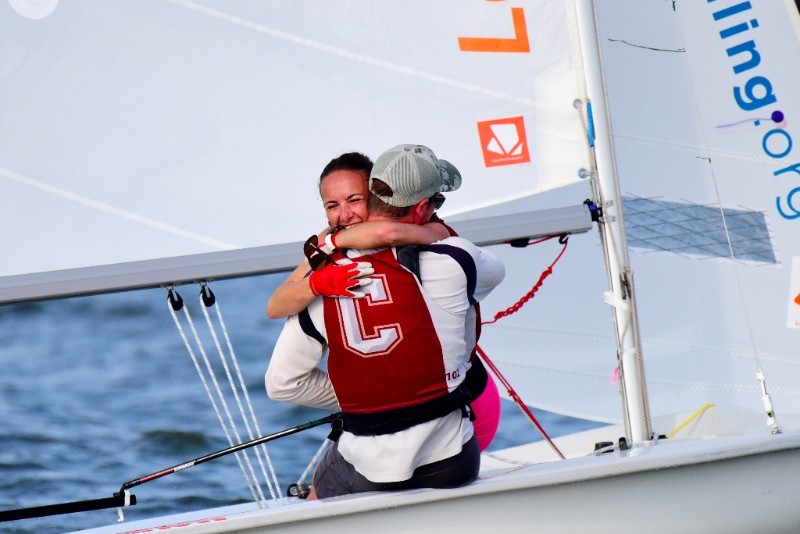 Two sailors wearing life vests embrace joyfully on a small sailboat, smiling with happiness. The water is visible in the background, and part of the sail shows the text 'ing.org.
