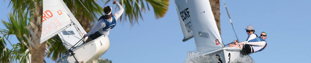 Two sailboats with crews wearing life vests are tilted sharply as they race on the water near palm trees under a clear blue sky.