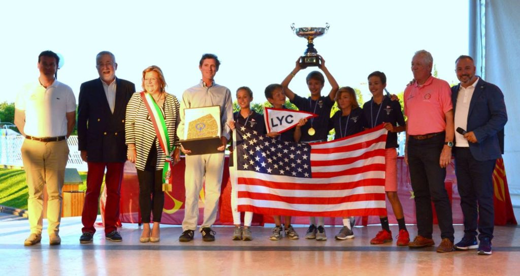 A group of people, including children and adults, stand together indoors. Some children hold a trophy and an American flag, while others hold medals and a framed plaque. Everyone is smiling and facing the camera.