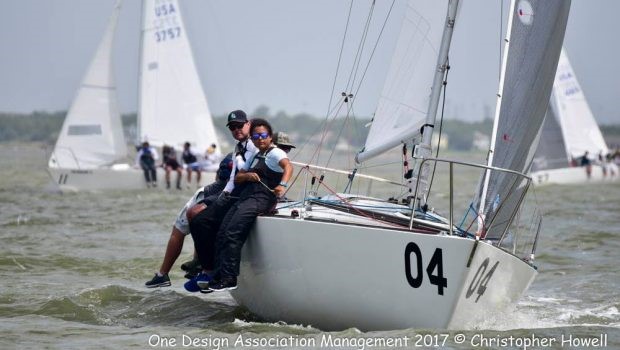 A sailboat marked '04' with four people onboard leans in the water during a sailing race, with other sailboats visible in the background. The crew is balancing on the edge, and the water appears choppy.