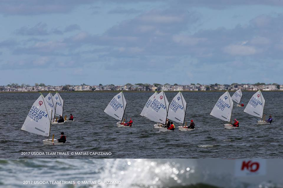A group of young sailors in small sailboats race across a choppy body of water near a distant shoreline under a partly cloudy sky. The sails display various numbers and symbols.