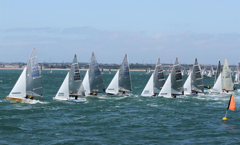 Several sailboats with white sails compete in a race on choppy blue water, heading toward an orange buoy; other boats and a distant shoreline with buildings are visible in the background.