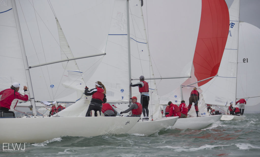 Several sailboats with crews in red jackets race closely together in choppy water, adjusting sails as they navigate. White and red sails billow in the wind under a cloudy sky.