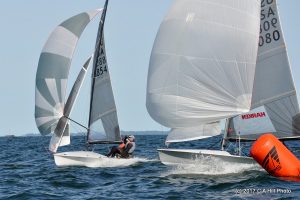 Two sailboats with white sails race closely around an orange buoy marker on open water, with sailors leaning to balance their boats under a clear blue sky.