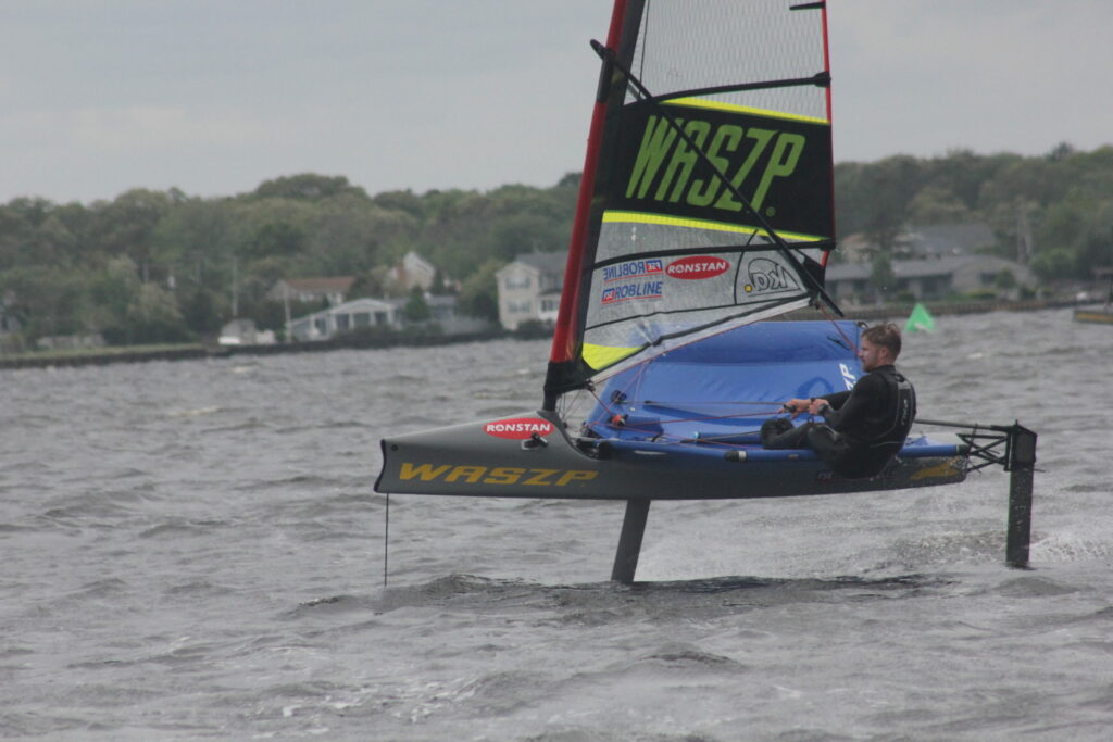 A person sails alone on a black and yellow hydrofoil dinghy labeled 'WASZP,' gliding above choppy water with houses, trees, and a cloudy sky in the background.