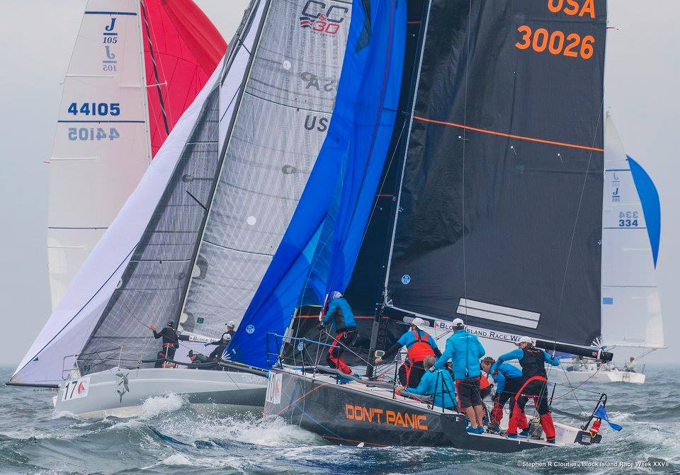 Several sailboats with colorful sails, including one labeled 'DON'T PANIC,' race closely together on the water. Crew members in bright clothing are actively managing sails under overcast skies.