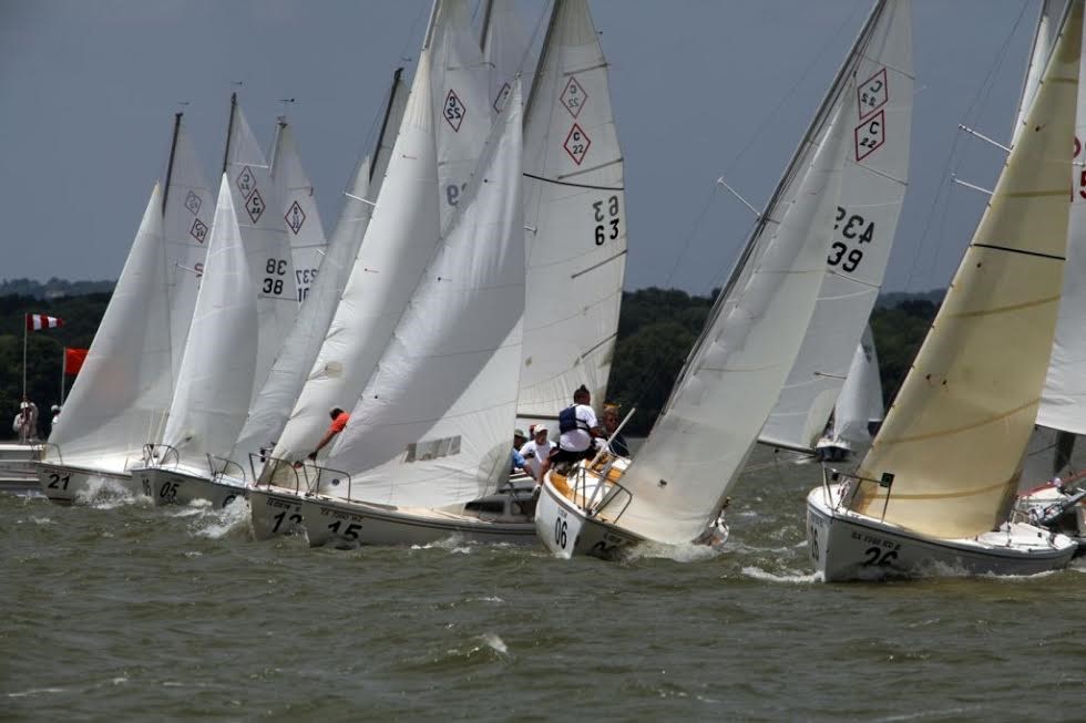 Several sailboats with numbered sails are racing closely together on choppy water under a cloudy sky, with trees visible in the background. Sailors maneuver the boats as they lean into the wind.