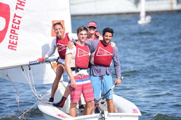 Four smiling people in red life vests stand on a small sailboat with "RESPECT" printed on the sail, posing for a photo on calm water on a sunny day.