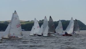 Six small sailboats with white sails are racing on a choppy body of water, with wooded hills and green fields in the background under a cloudy sky.