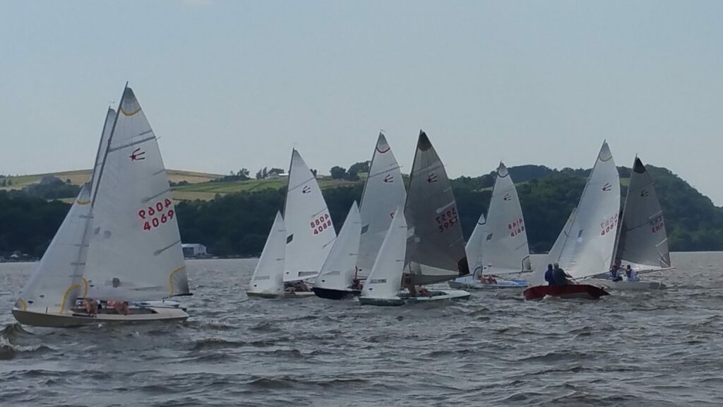 Six small sailboats with white sails are racing on a choppy body of water, with wooded hills and green fields in the background under a cloudy sky.