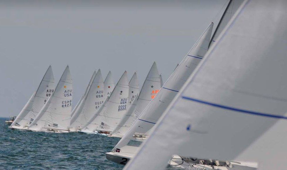 A row of sailboats with marked sails races closely together on the water under a cloudy sky, with their white sails angled uniformly in the wind.