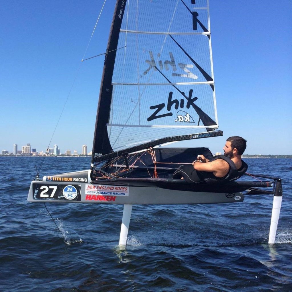 A man sails alone in a small hydrofoil sailboat with the number 27, gliding above the water on foils. City buildings and a clear blue sky are visible in the background.