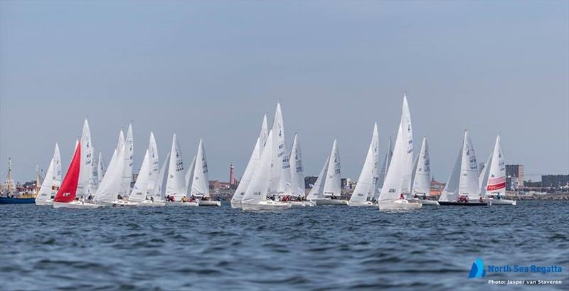 A group of sailboats with white sails participate in a regatta on calm water, with a cityscape and lighthouse visible in the background. One sailboat has a distinct red sail. North Sea Regatta logo is in the corner.