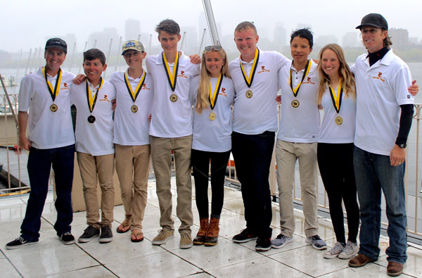 Eight young people and one adult, all wearing white polo shirts and medals, stand arm-in-arm on a dock with boats and a foggy city skyline in the background, smiling for the camera.