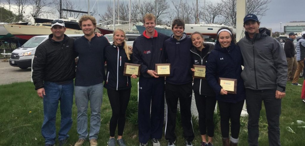 A group of seven people stands outdoors in front of sailboats, smiling and holding plaques, dressed warmly in jackets and athletic wear, suggesting they have won an award at a sailing event.