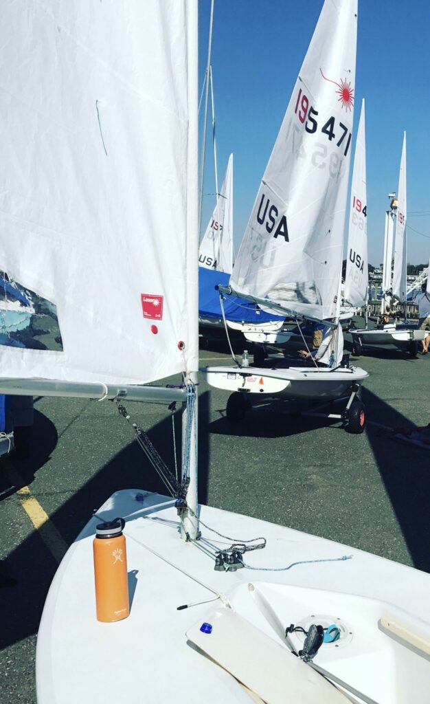 Several small sailboats with white sails are on land, with one close in the foreground showing deck details and an orange water bottle. Other sailboats and clear blue sky are visible in the background.