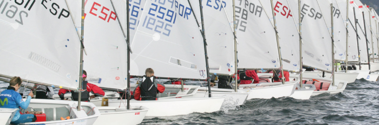 A row of young sailors in small sailboats compete in a regatta, each boat with white sails displaying numbers and country codes, sailing closely together on choppy water.