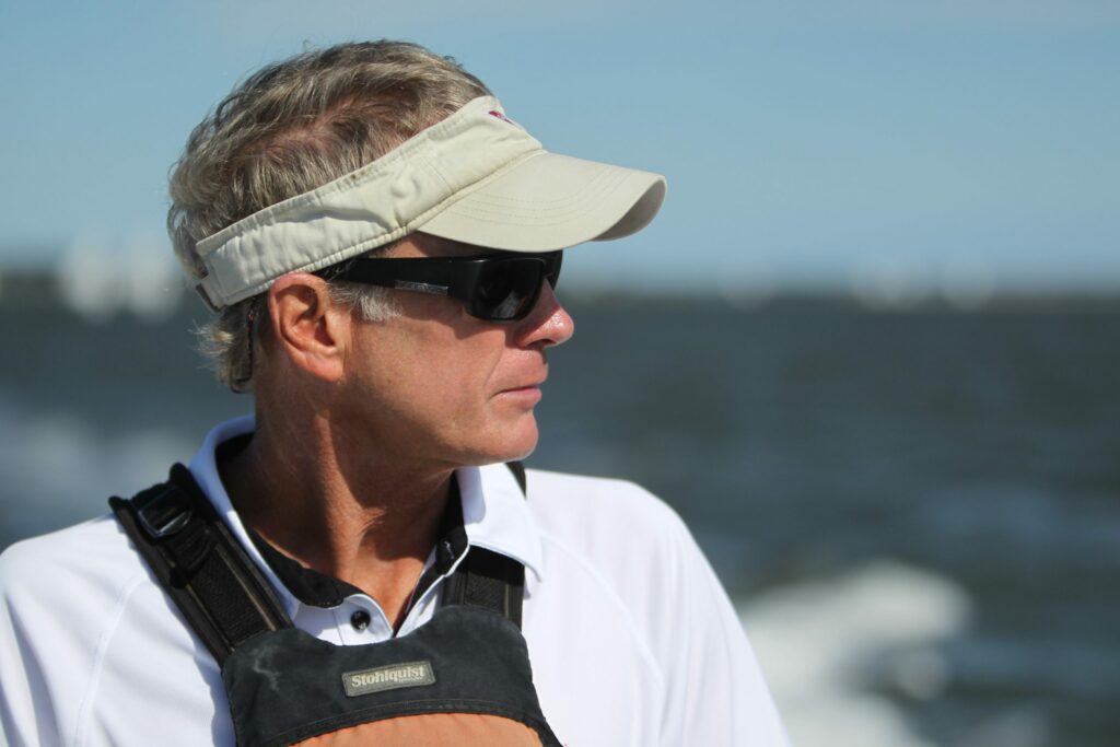 A man wearing a light-colored visor, dark sunglasses, and a white shirt stands outdoors near water, looking to the side. The background shows blurred waves and a blue sky.