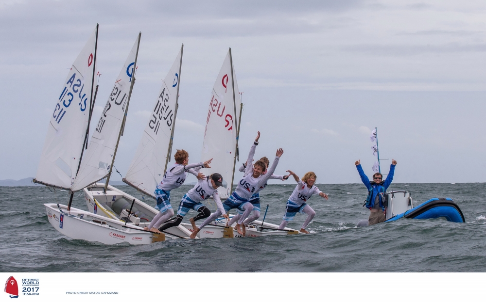 Four young sailors in matching uniforms celebrate and jump off their sailboat into the water, while a coach in a support boat cheers nearby. Waves and cloudy skies are visible in the background.