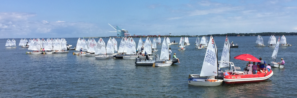 A large group of small sailboats with white sails gather on calm water near the shore. People are visible on the boats and on a red-and-white motorboat, with an industrial area and trees in the background under a blue sky.