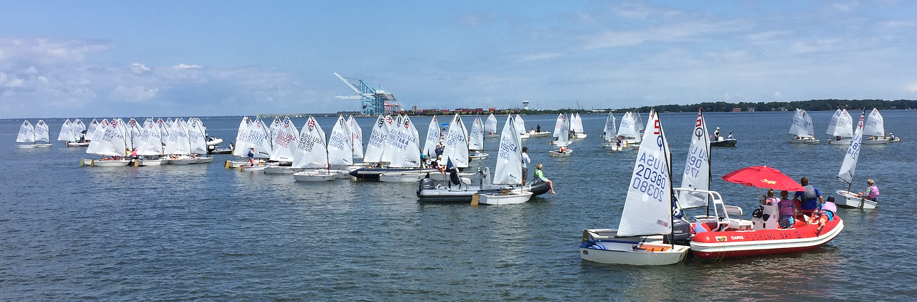 A large group of small sailboats with white sails gather on calm water near the shore. People are visible on the boats and on a red-and-white motorboat, with an industrial area and trees in the background under a blue sky.