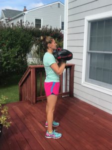 A woman stands on a wooden deck holding a weighted sandbag at chest height, facing a gray house with white trim. She wears a light blue shirt, pink shorts, and colorful sneakers.