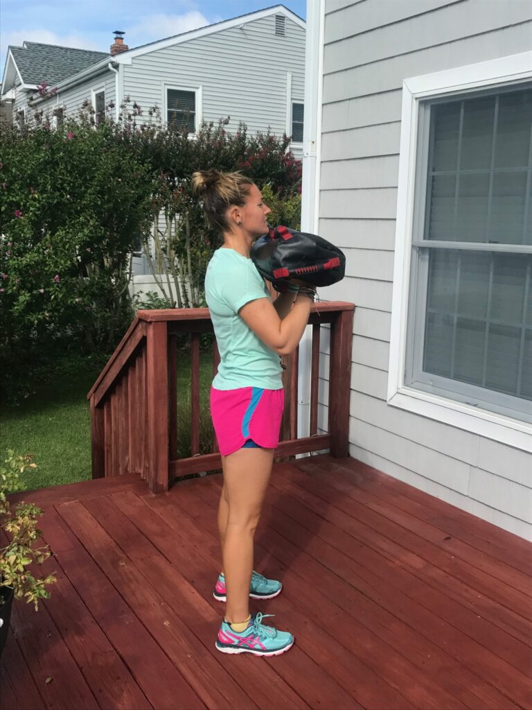 A woman stands on a wooden deck holding a weighted sandbag at chest height, facing a gray house with white trim. She wears a light blue shirt, pink shorts, and colorful sneakers.