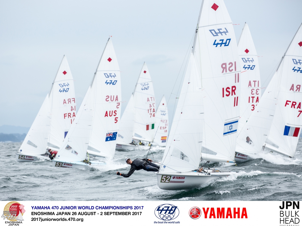 Several sailboats with competitors race closely together on the water during the 2017 Yamaha 470 Junior World Championships in Enoshima, Japan. Event logos and sponsor names are visible at the bottom of the image.