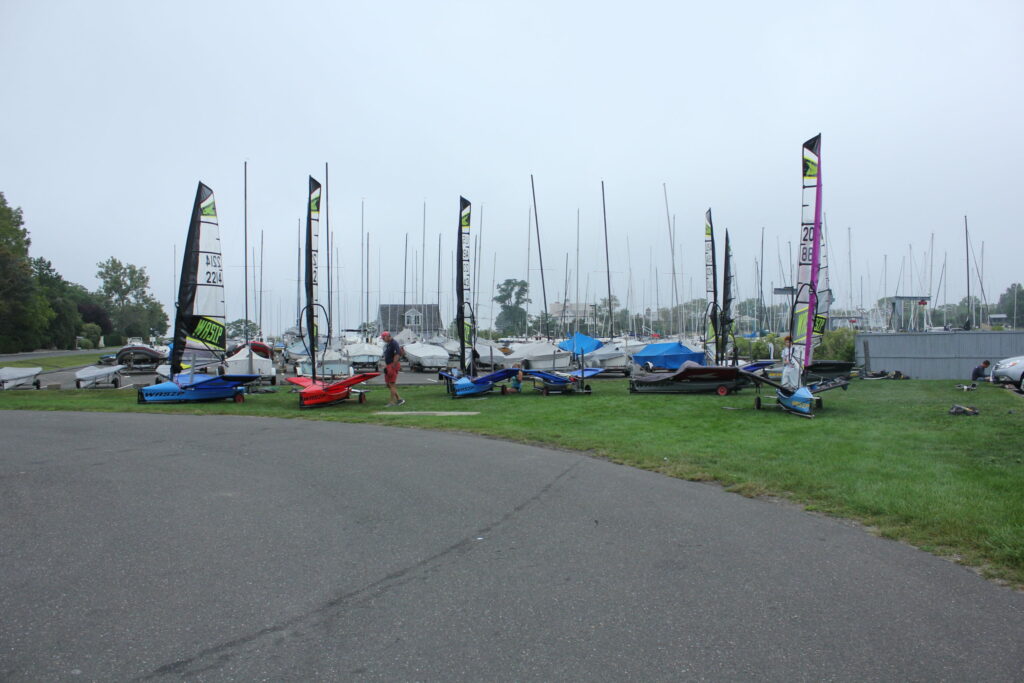 Several small sailboats with colorful sails are parked on grass near a marina, with many sailboat masts visible in the background and a person walking among the boats on a cloudy day.