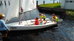 Three children wearing life jackets are getting ready to sail a small white sailboat near a dock, with an adult standing in the shallow water beside them, offering guidance.