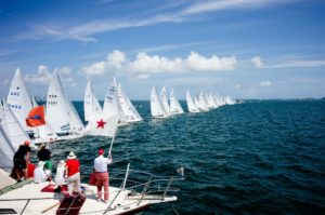 A line of sailboats with white sails race across blue water under a partly cloudy sky, as people on a nearby boat watch and wave flags.