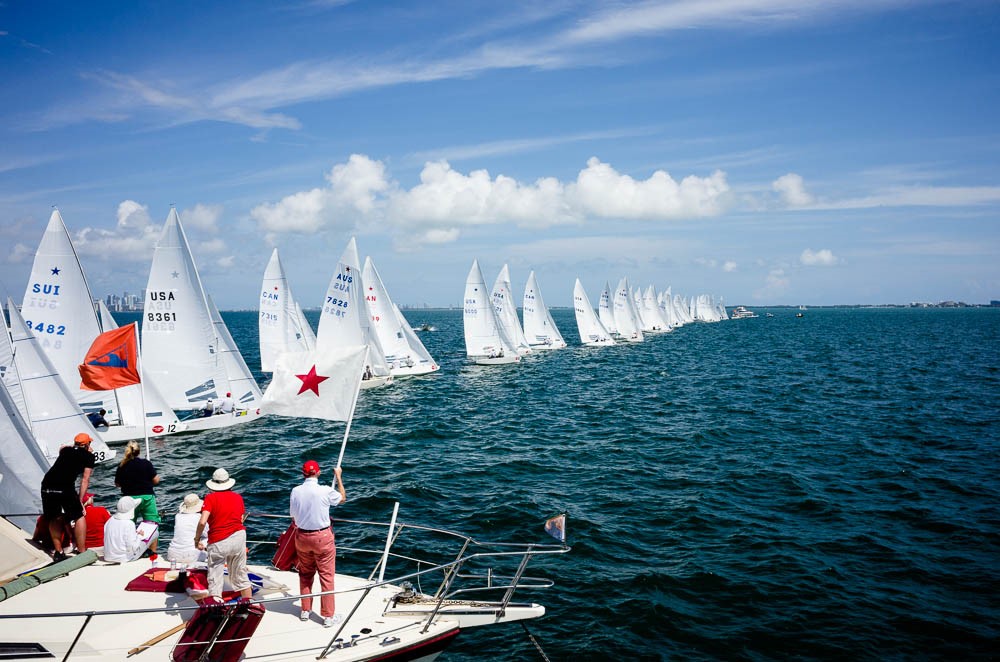 A line of sailboats with white sails race across blue water under a partly cloudy sky, as people on a nearby boat watch and wave flags.