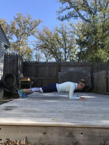A person in athletic clothing performs a forearm plank on a wooden deck outside, with a fence, trees, and grill visible in the background on a sunny day.